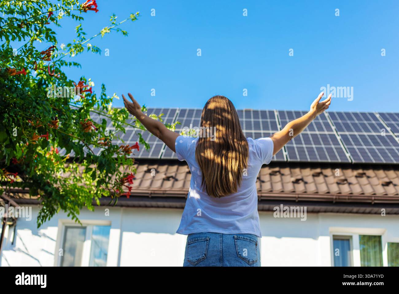 Donna con le braccia alzate di fronte a una casa con pannelli solari e tromba in fiore. Foto Stock