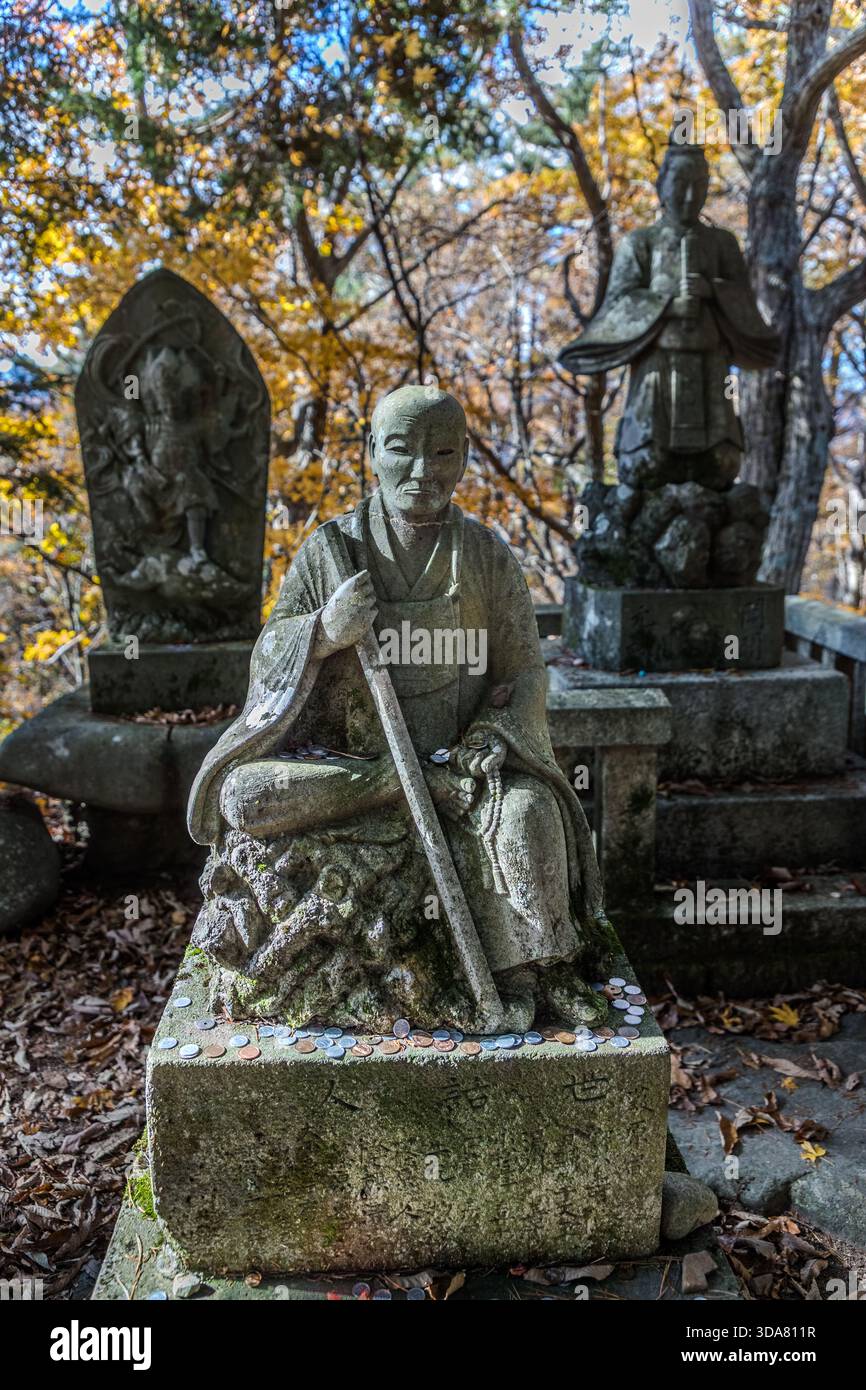 Statua di un monaco seduto con il personale di un pellegrino e offerte di monete al passo di Toriitoge, Nakasendo, valle di Kiso, prefettura di Nagano, Giappone; altre statue sono visibili sullo sfondo Foto Stock