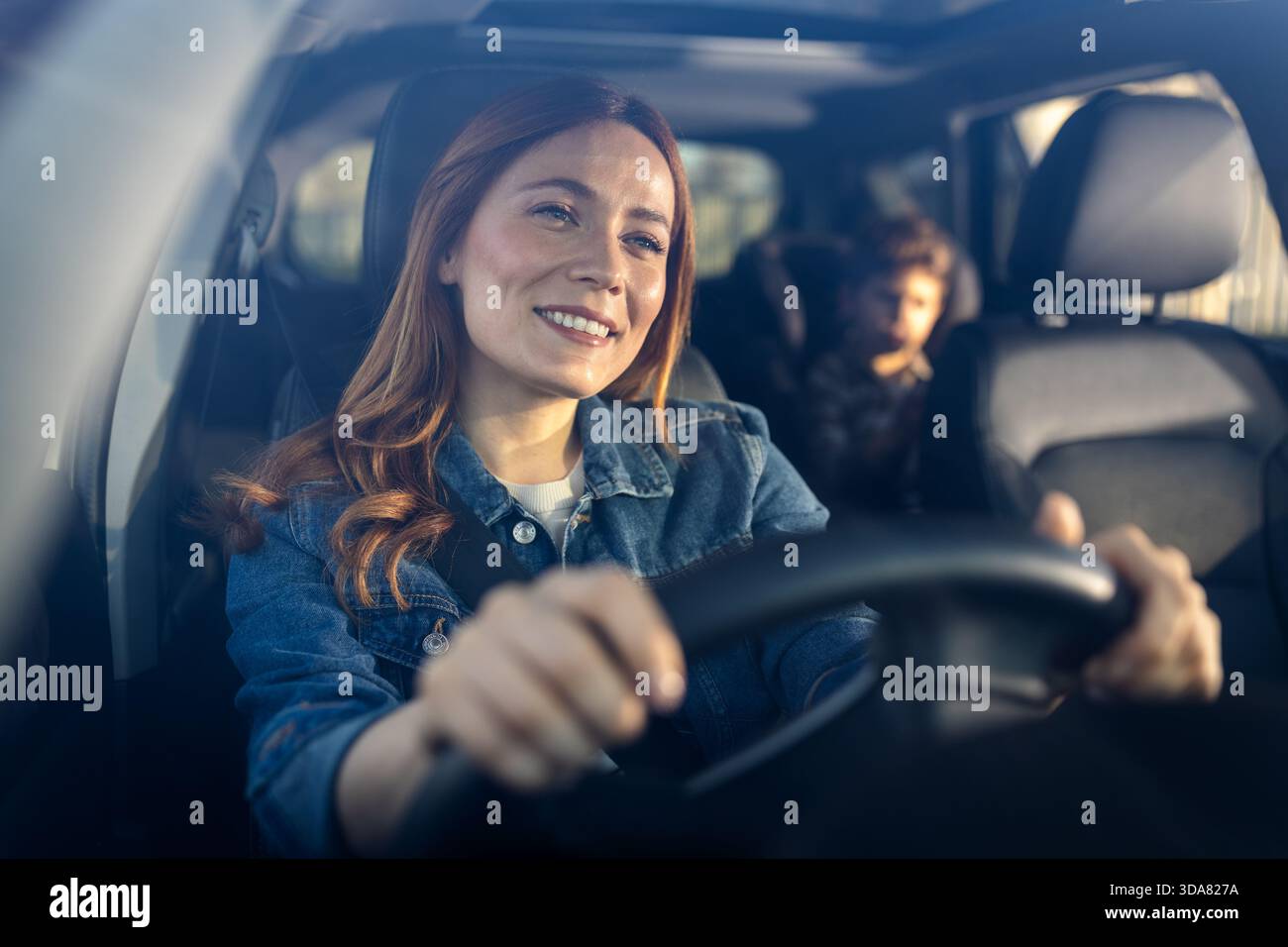 Donna sorridente durante la guida dell'auto, con bambino seduto comodamente sul sedile posteriore. La luce del sole illumina il loro viaggio Foto Stock