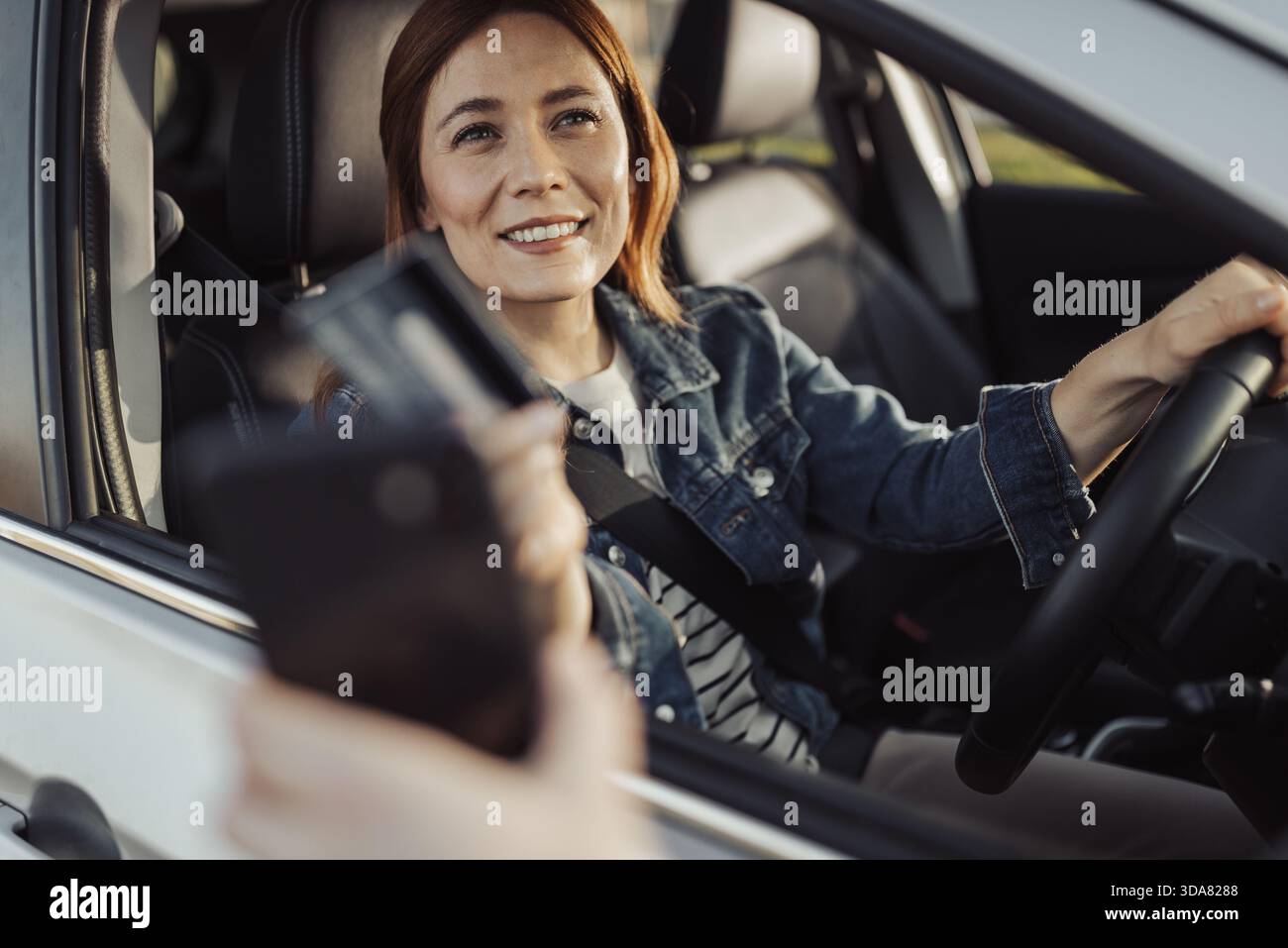 Una donna siede in macchina sorridendo mentre si prepara a pagare al drive-thru. La scena mostra un pomeriggio impegnativo in un fast food. Foto Stock