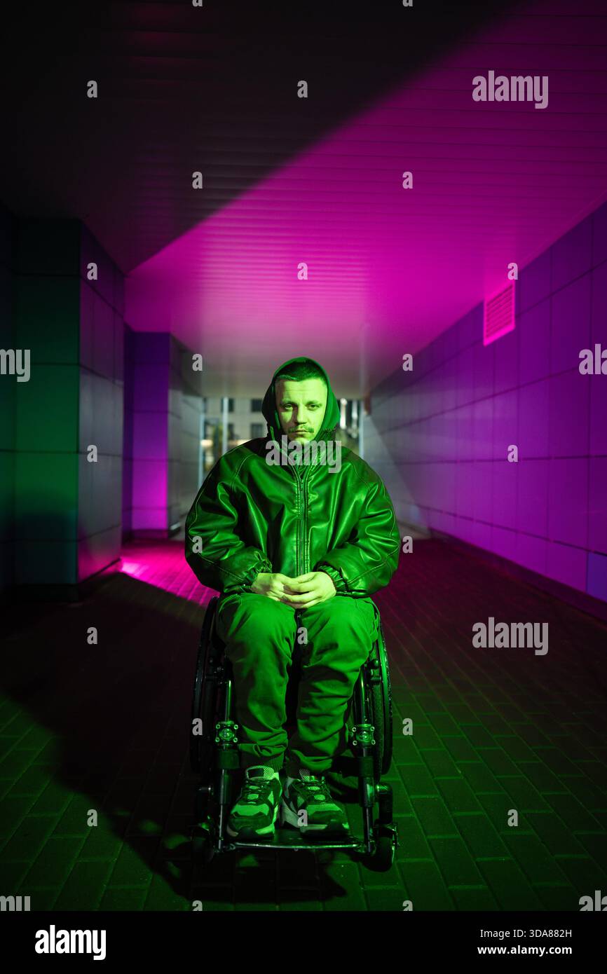 Giovane uomo disabile pensivo in sedia a rotelle che cammina da solo, seduto in una luce rgb al neon a colori guardando la macchina fotografica con un'espressione triste e stanca. Uomo con disabilità che mostra segni di stato mentale pesante e apatia || modello rilasciato Foto Stock