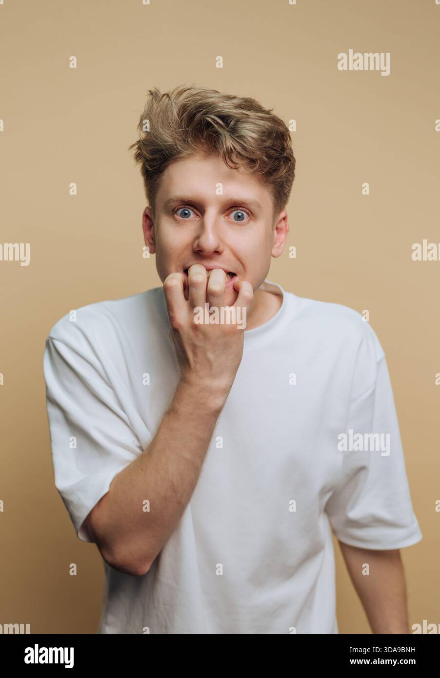Un giovane con i capelli duri che sembra ansioso, che si morde le unghie, su uno sfondo beige Foto Stock
