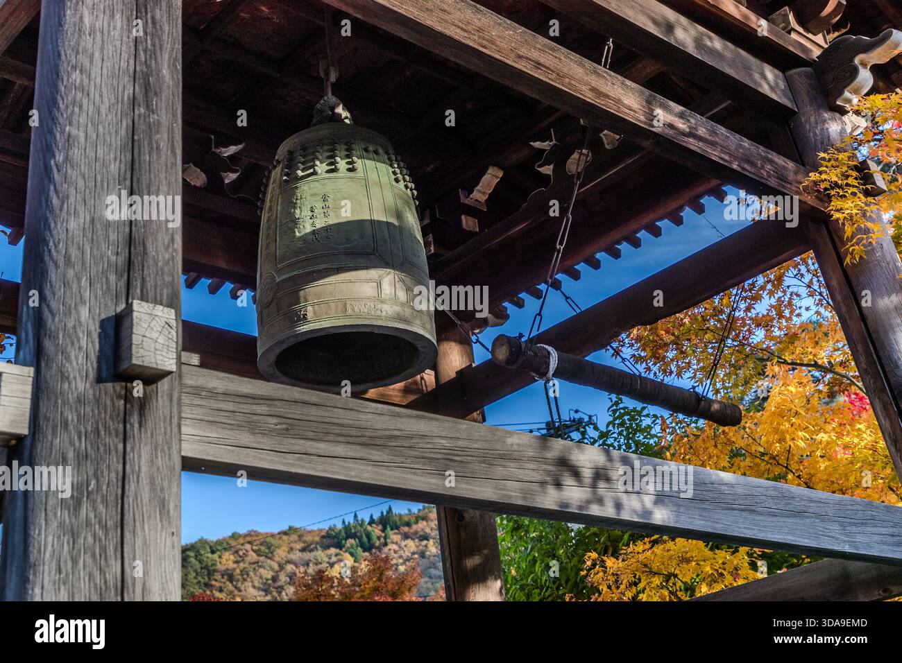 Una campana del tempio in bronzo (bonshō) con un'iscrizione incisa è appesa nel padiglione della campana in legno, con alberi nelle tonalità autunnali dell'oro e del rosso sullo sfondo, Kiso, prefettura di Nagano, Giappone Foto Stock