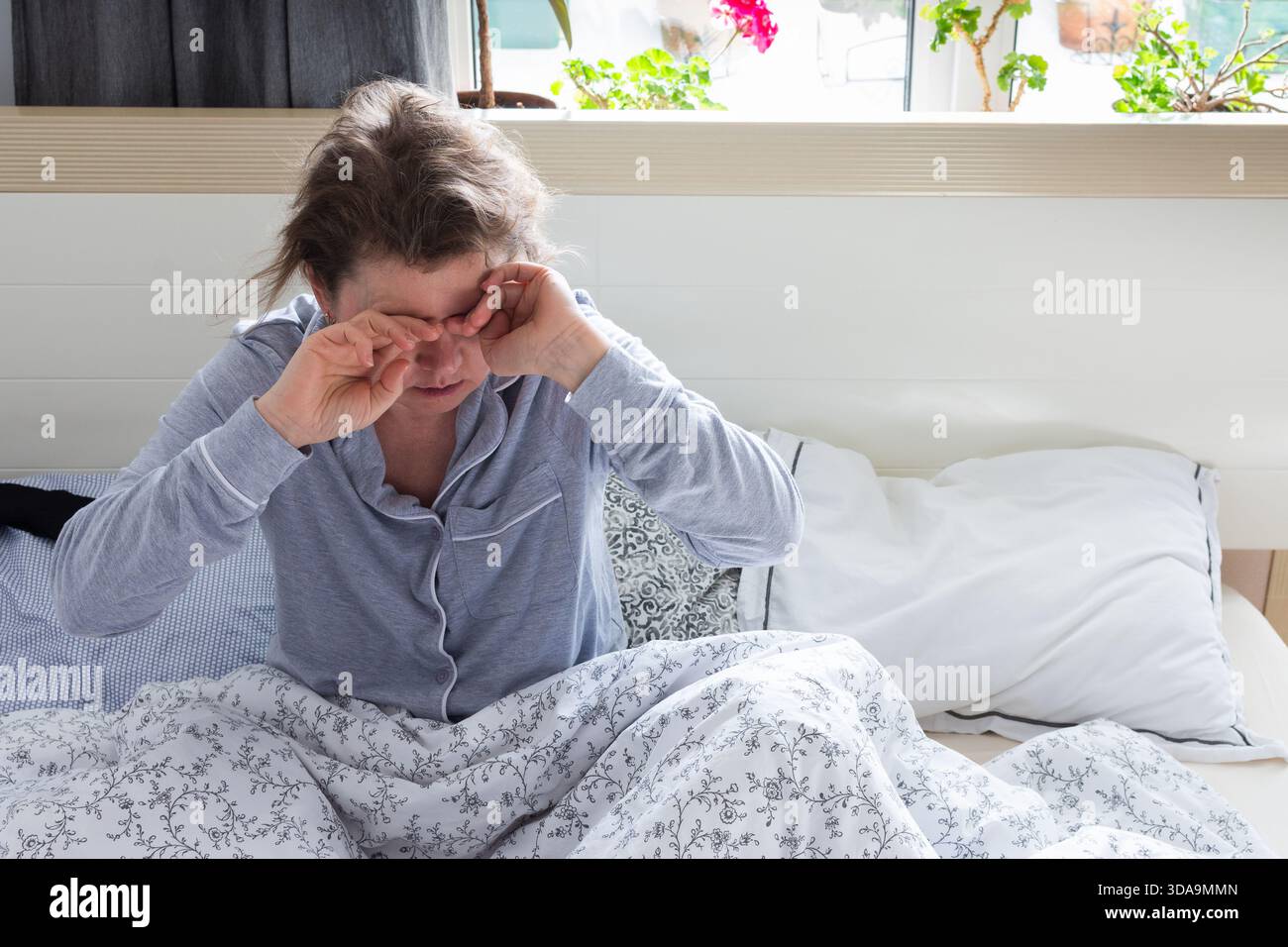 Una donna di 50 anni appare stanca e angosciata mentre si siede a letto. Si strofina gli occhi, indicando una mancanza di sonno, mentre è circondata da un letto morbido Foto Stock