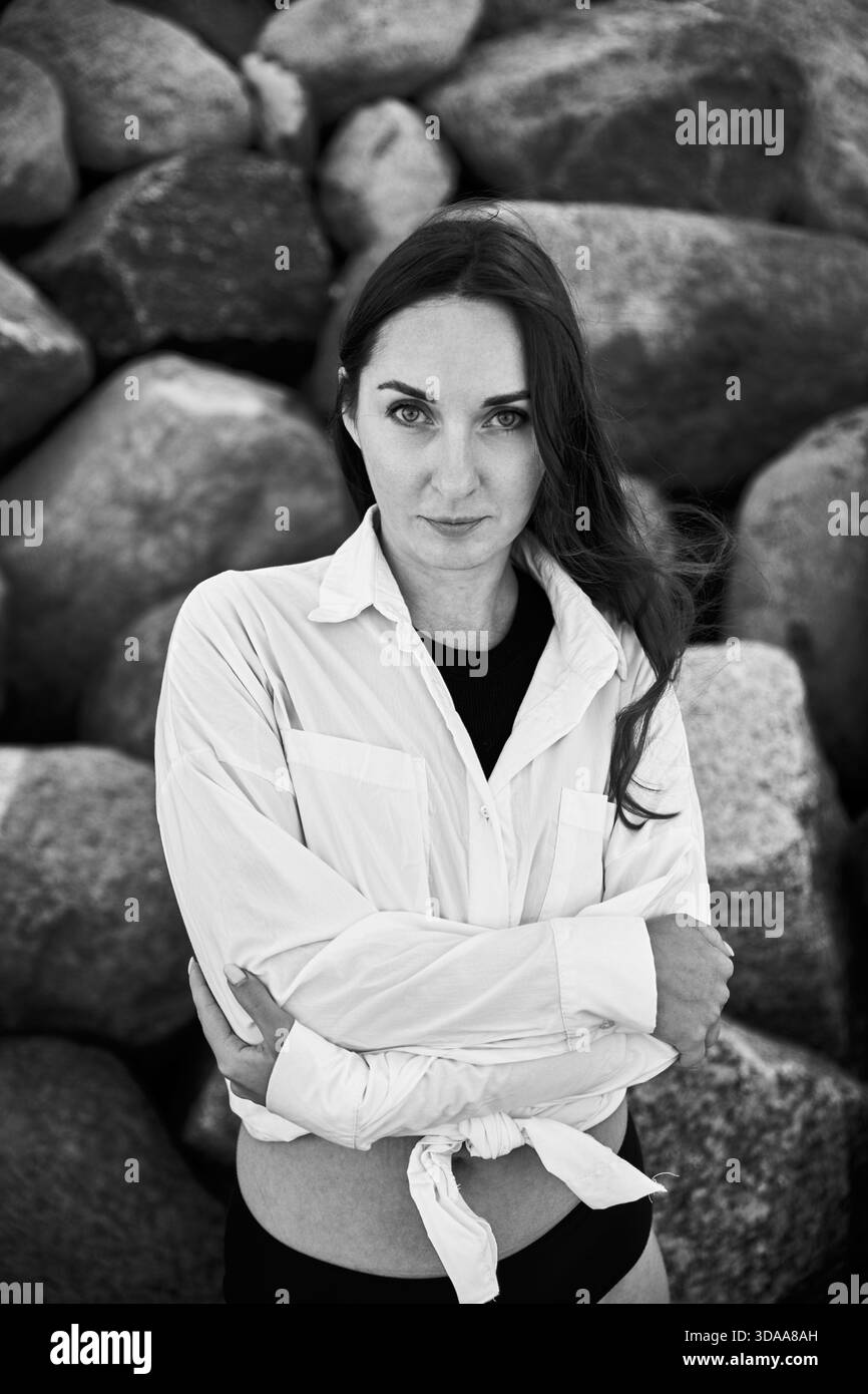 Donna adulta con lunghi capelli scuri in piedi con sicurezza contro grandi rocce. Il tono bianco e nero enfatizza l'espressione del viso e lo sfondo testurizzato Foto Stock