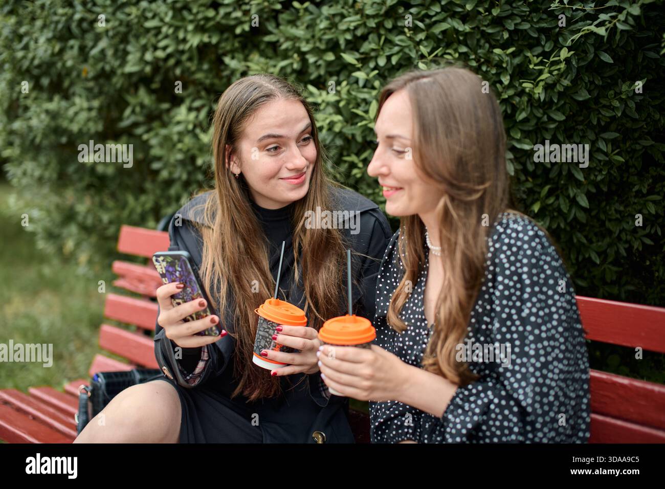 Due giovani donne adulte con capelli lunghi siedono sulla panchina rossa all'aperto, tenendo tazze da caffè da asporto e sorridendo, impegnate in pettegolezzi amichevoli con siepe verde Foto Stock