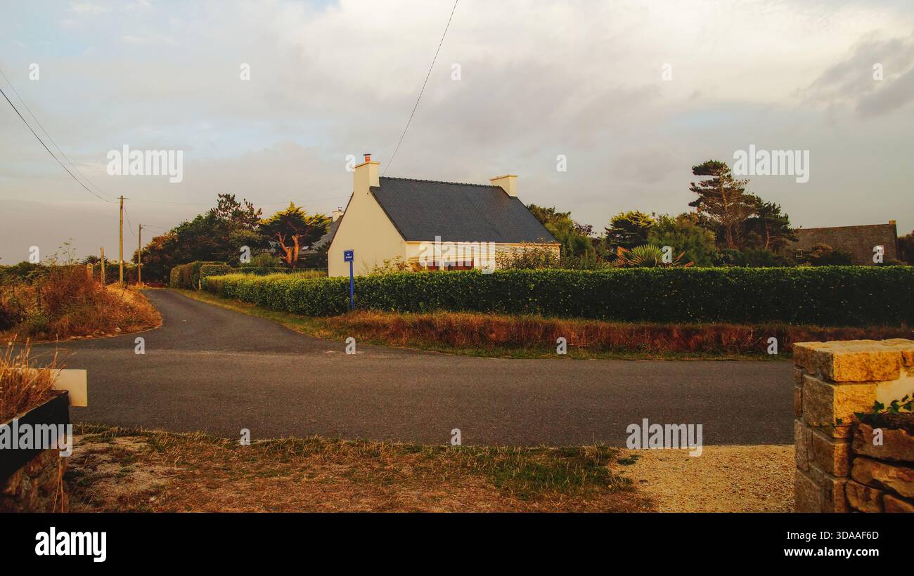 Casa solitaria accanto a una strada di campagna vuota al crepuscolo, circondata da verde e luce soffusa. Una tranquilla scena rurale con un'atmosfera serena. Foto Stock