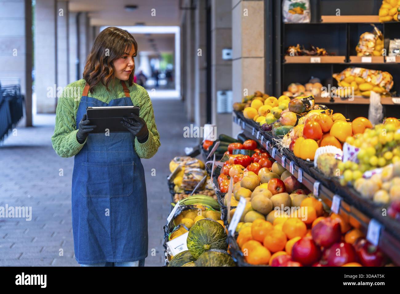 Droghiere donna che utilizza un tablet digitale per gestire l'inventario e i prodotti freschi presso il suo stand di frutta e verdura all'aperto, mettendo in evidenza la tecnologia moderna Foto Stock