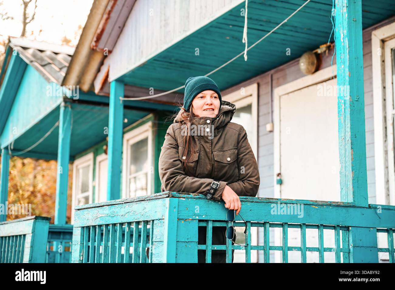 Donna russa in piedi sul portico della sua casa rurale in legno, indossando abiti caldi e cappello. Accogliente vita da villaggio in autunno, inverno o inizio primavera. Foto Stock