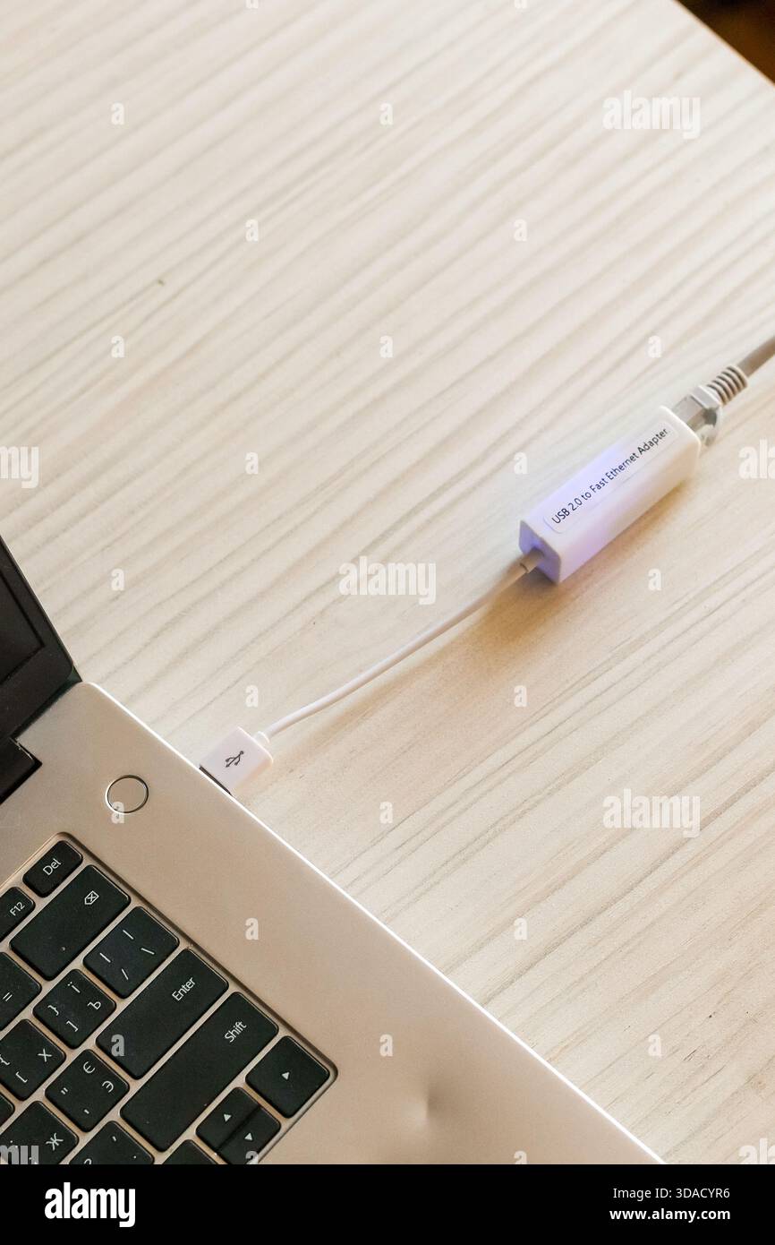 Immagine dettagliata di un adattatore Ethernet USB bianco con cavo di rete collegato appoggiato su una scrivania luminosa vicino al bordo di un laptop, evidenziando l'hardwa della connettività Foto Stock
