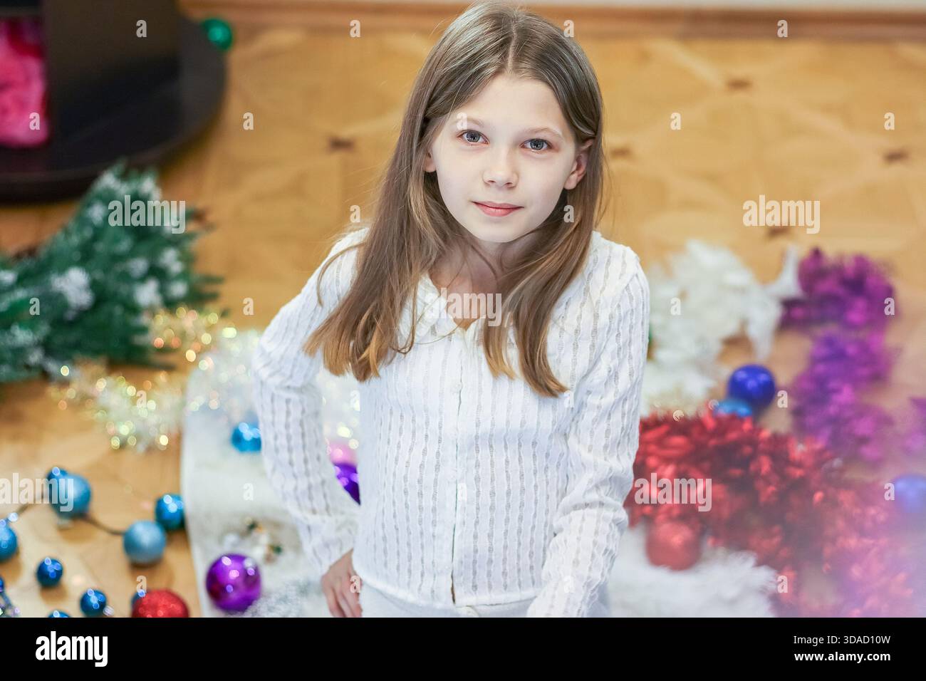 Foto top-down di preadolescente con mani sui fianchi in abito bianco tra ornamenti natalizi, tinsel e rami di pino sul pavimento. Foto Stock