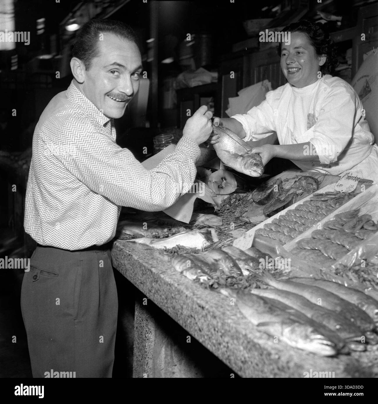 Giocatore di rugby al mercato del pesce (Place Victor-Hugo, Tolosa, Francia, 23–24 agosto 1956): Vincent Cantoni, centro a tre quarti dell'Olympique XIII di Tolosa, si erge di fronte a una bancarella di pesci al mercato Victor-Hugo come venditore femminile gli consegna un pesce; gira la testa verso la macchina fotografica, catturando un momento vivace della vita quotidiana del mercato e della cultura delle celebrità sportive nella Francia della metà degli anni '1950. Foto Stock