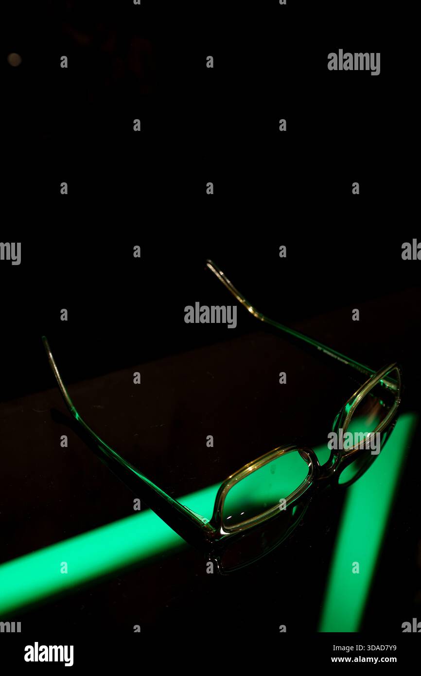 Un paio di occhiali poggiati su una superficie nera lucida illuminata da una forte luce verde al neon. Foto Stock