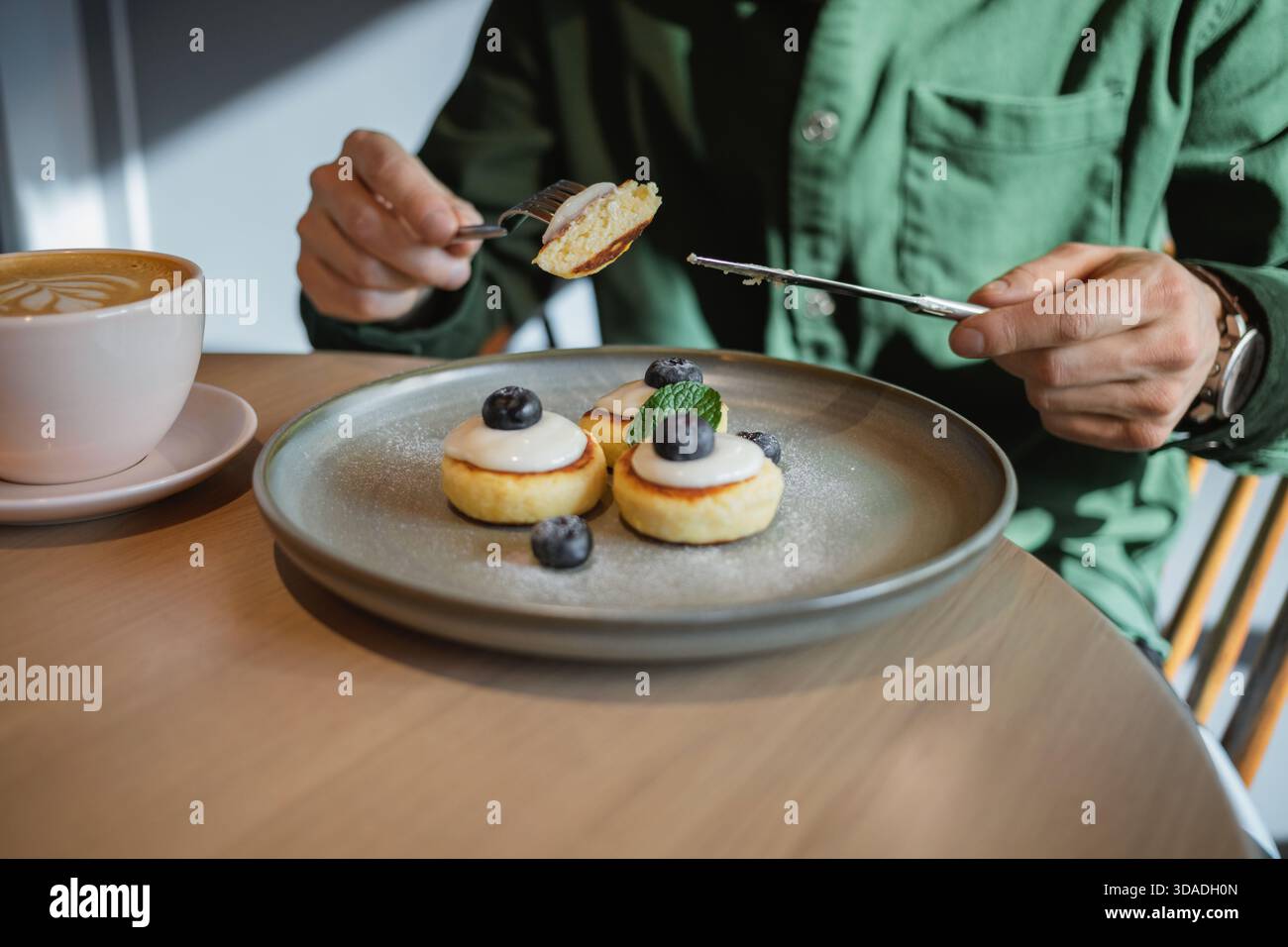 Primo piano di un uomo seduto a un tavolo che mangia Mini Pancakes con yogurt fresco, mirtilli, menta e un cappuccino Foto Stock