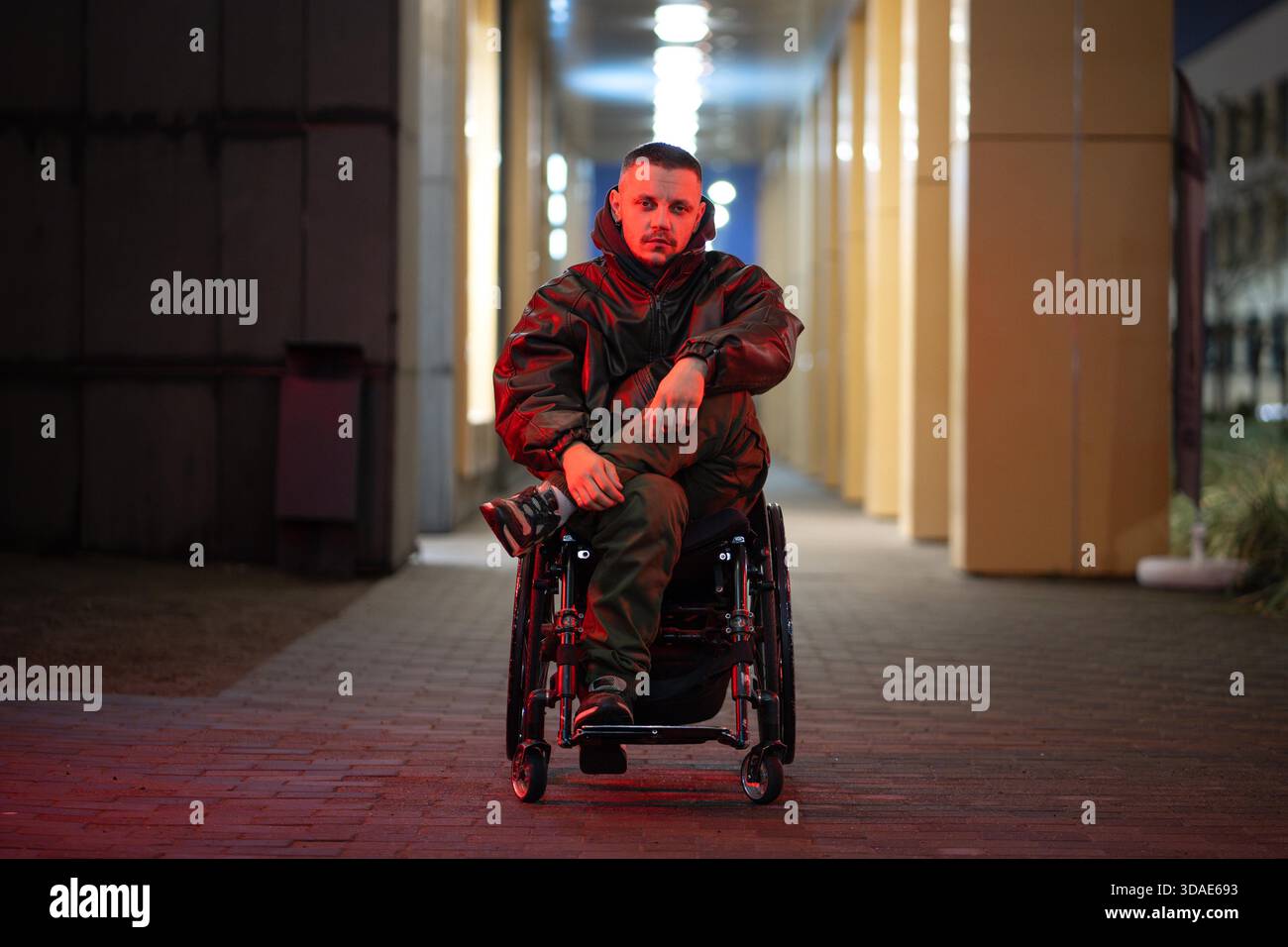 Giovane disabile in sedia a rotelle con un'espressione sicura durante una passeggiata notturna in città, che posa guardando la fotocamera. Uomo con disabilità che mantiene attivo lo stato mentale e la motivazione nonostante l'immobilità. || modello rilasciato Foto Stock