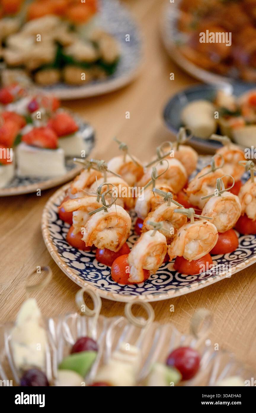 Tartine di gamberi sugli spiedini con pomodori ciliegini su un piatto decorativo su un tavolo di legno. Un buffet di antipasti con gamberi tigre, pomodori e frutta c Foto Stock