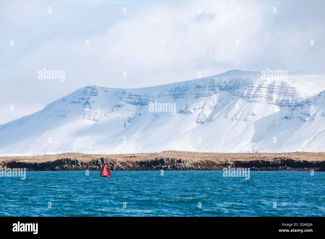 Un tranquillo paesaggio marino è caratterizzato da una boa rossa che galleggia in un mare blu profondo, con una catena montuosa coperta di neve e una costa rocciosa che crea un freddo, i Foto Stock