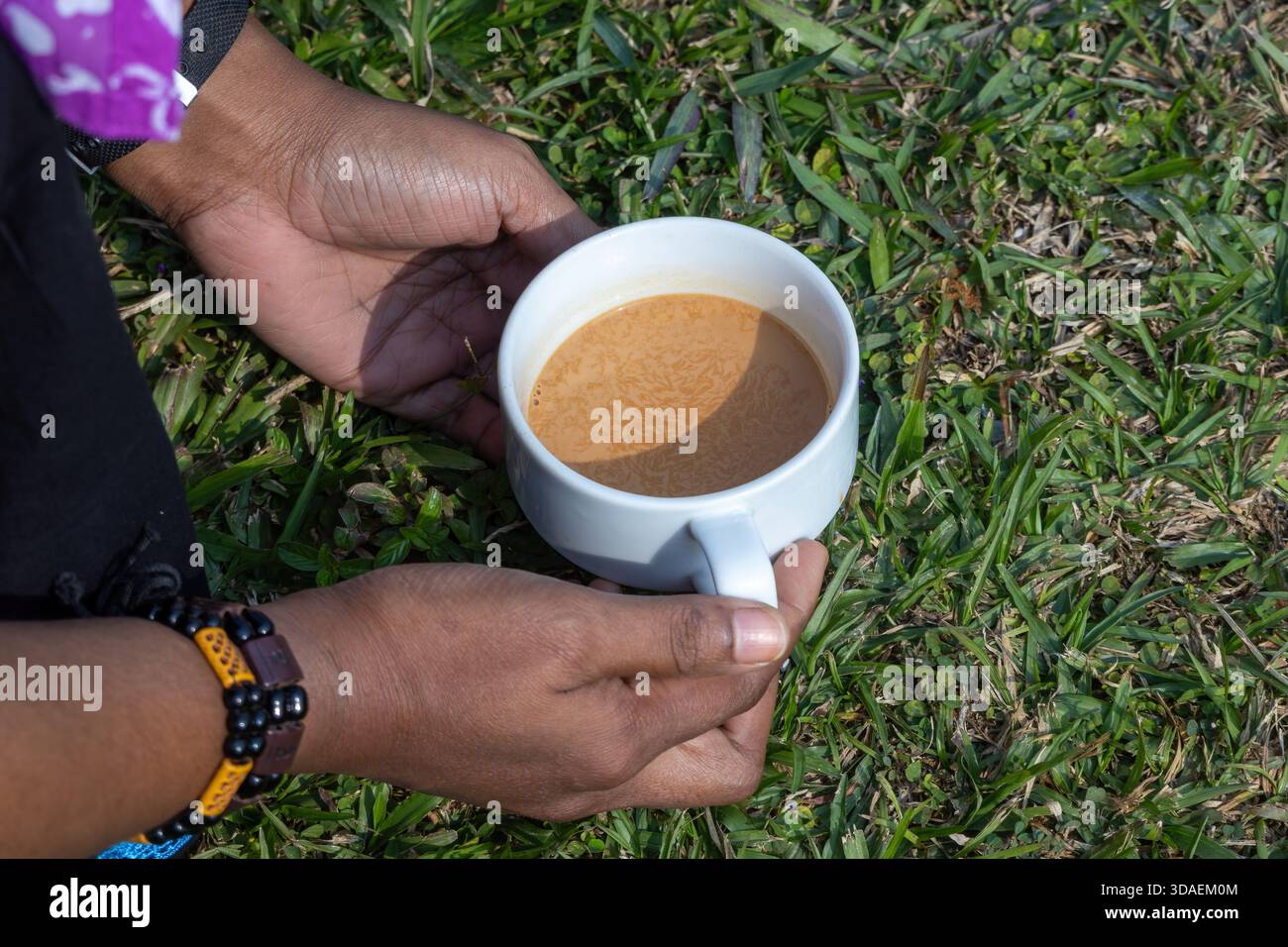Primo piano di una mano femminile che tiene in mano una tazza bianca di tè al latte caldo (Chai) mentre si siede su una lussureggiante erba verde all'aperto. Momenti rilassanti e informali Foto Stock