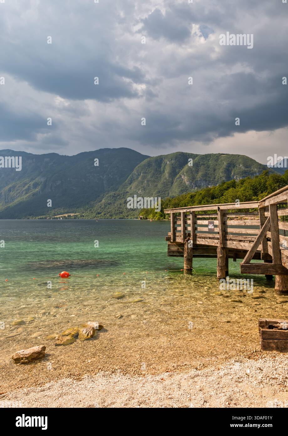Il molo in legno si estende nel lago turchese con boa rossa, riva di ciottoli e verdi montagne sotto il cielo spettacolare. Foto Stock