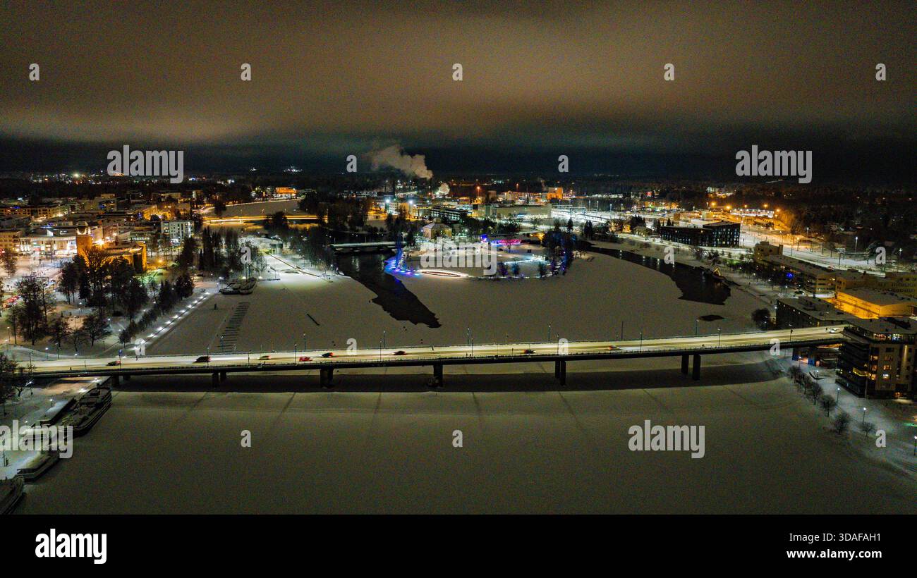 Veduta aerea di un ponte illuminato sul ghiacciato fiume Pielisjoki, in contrasto con le acque scure e le luci della città che brillano in lontananza, Joe Foto Stock