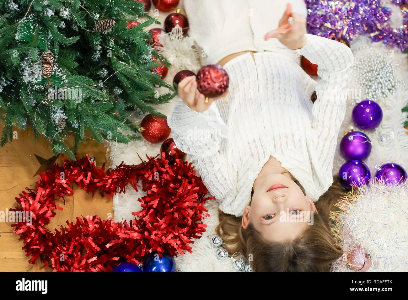 Il bambino in un outfit bianco testurizzato afferra la sfera festosa su un morbido tappeto circondato da tinsel, palle e rami di pino Foto Stock