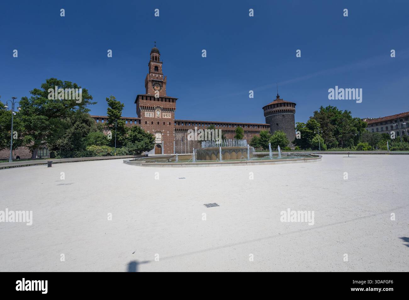 Un castello europeo illuminato dal sole presenta un'alta torre accanto ad una fontana circolare con getti d'acqua. Alberi lussureggianti e pareti in mattoni fanno da cornice a una scena culturale senza tempo della città Foto Stock