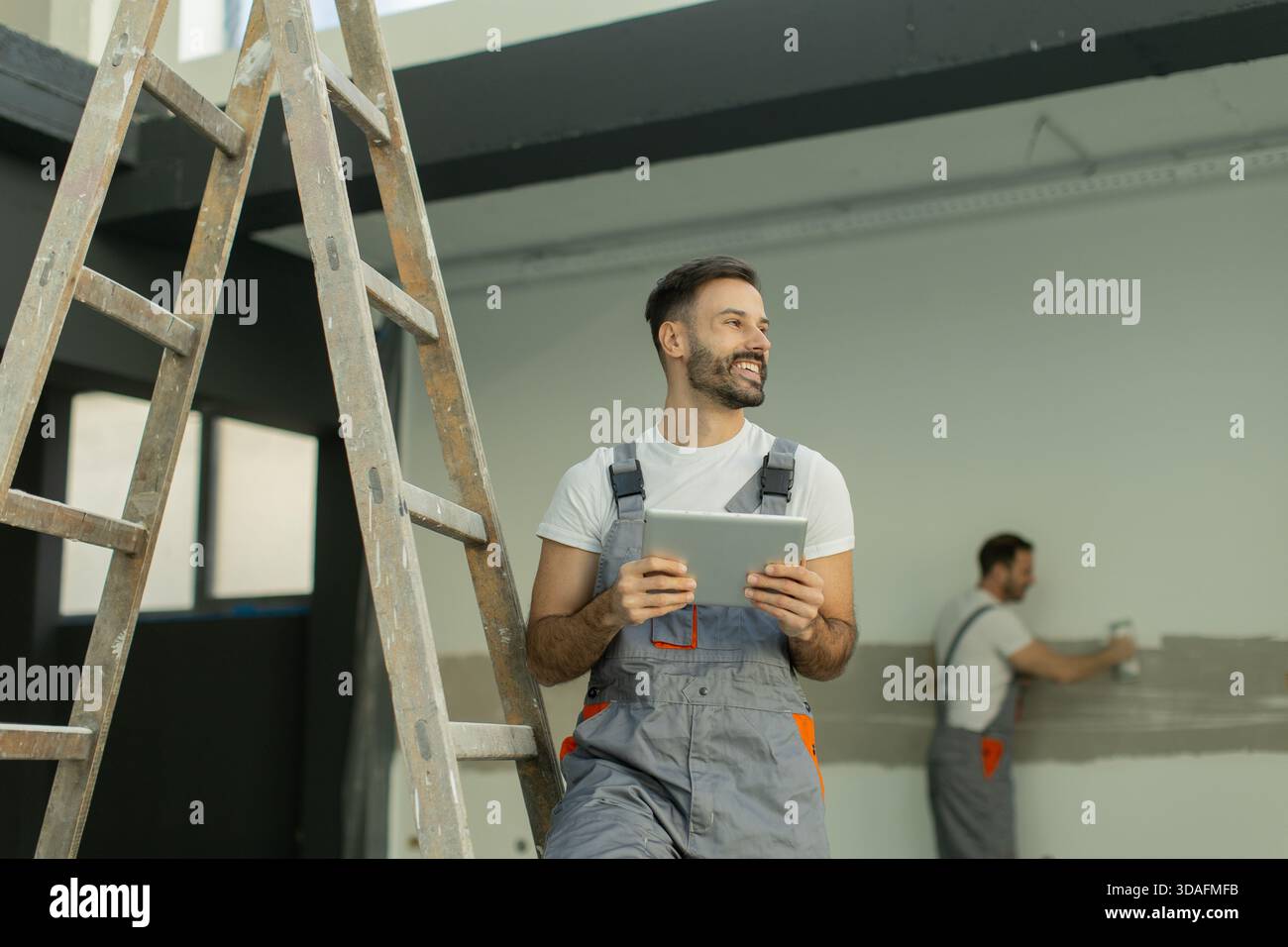 Due lavoratori sono impegnati in un progetto di ristrutturazione. Uno regge un tablet, sorride, mentre l'altro dipinge un muro. Sono in una spaziosa area interna durante Foto Stock