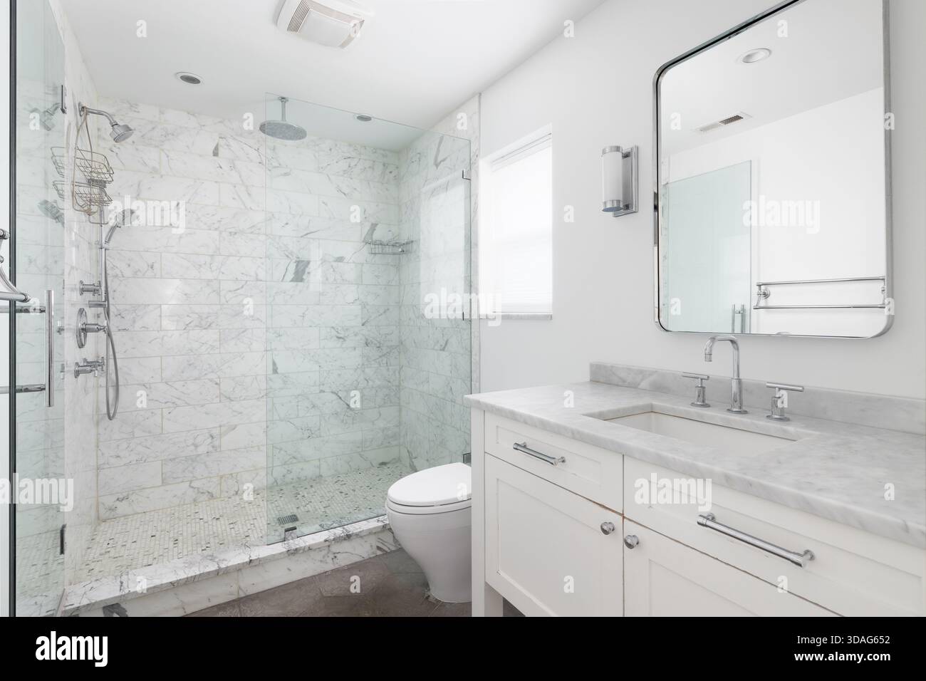 Un bagno bianco luminoso e moderno caratterizzato da una spaziosa cabina doccia in vetro con piastrelle in marmo e un piano lavabo bianco con ripiano in marmo. Foto Stock