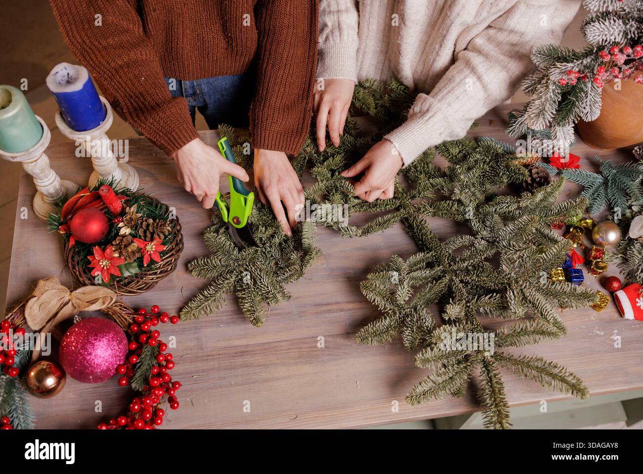 Vista ravvicinata di donne che tagliano rami sempreverdi con forbici da potatura mentre preparano decorazioni natalizie fatte a mano su un tavolo di legno. Foto Stock