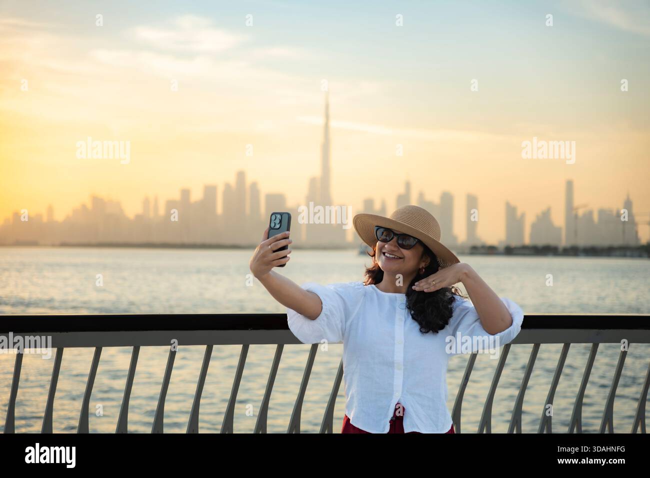 Donna turistica che scatta selfie con lo skyline di Dubai e l'iconico Burj Khalifa al tramonto, indossa un cappello con una camicia bianca, vacanze invernali e viaggi Foto Stock