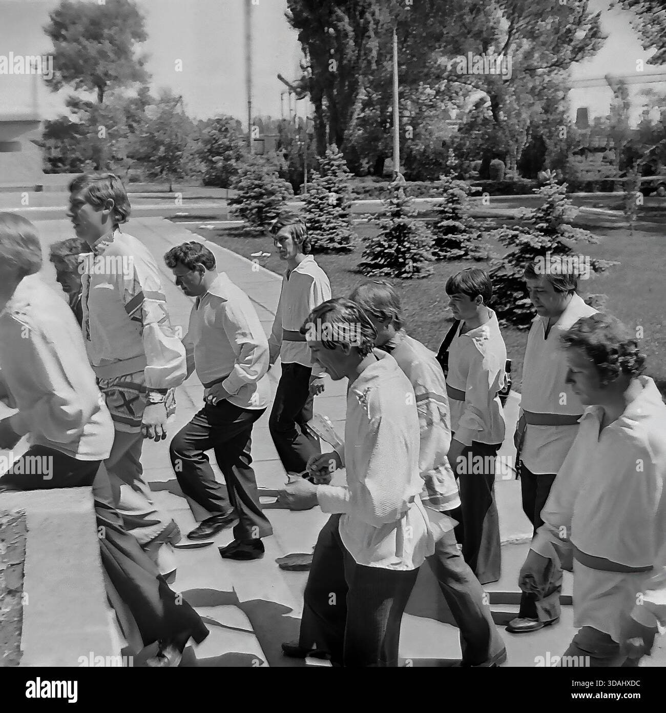 Regione di Donetsk, Ucraina, URSS. Maggio 1979. Un'autentica fotografia d'archivio in bianco e nero raffigurante i membri di un gruppo folk maschile che camminano attraverso un parco durante un festival d'arte dilettante del quartiere. Il gruppo di uomini è vestito con camicie di raso stilizzate (kosovorotkas) con ricami e fasce. Camminano davanti a alberi di abete blu vicino a un centro culturale, uno che porta un tamburello. Questa scena cattura il vibrante movimento artistico e l'atmosfera festosa del tranquillo Donbass. Foto Stock