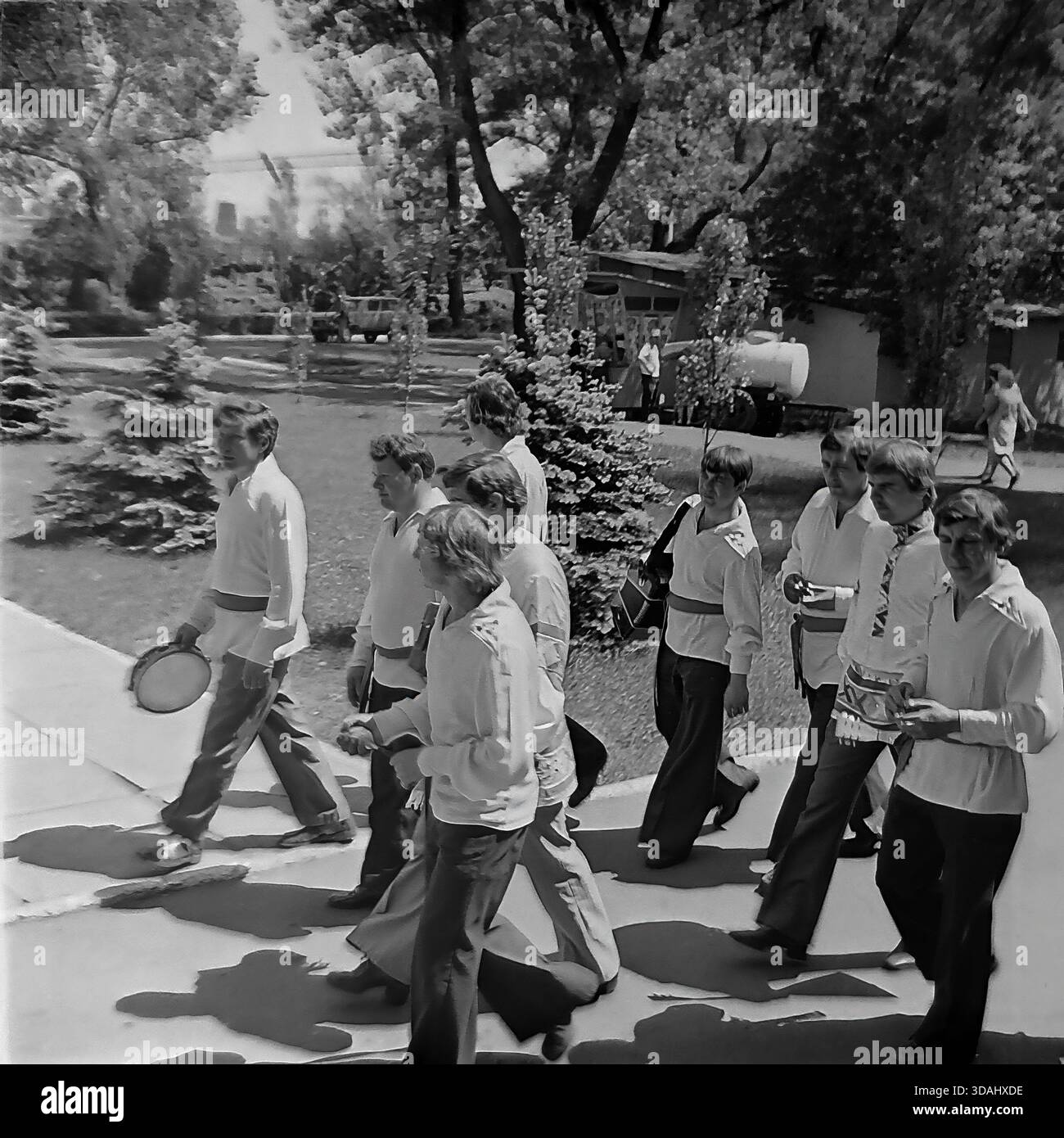 Regione di Donetsk, Ucraina, URSS. Maggio 1979. Un'autentica fotografia d'archivio in bianco e nero raffigurante i membri di un gruppo folk maschile che camminano attraverso un parco durante un festival d'arte dilettante del quartiere. Il gruppo di uomini è vestito con camicie di raso stilizzate (kosovorotkas) con ricami e fasce. Camminano davanti a alberi di abete blu vicino a un centro culturale, uno che porta un tamburello. Questa scena cattura il vibrante movimento artistico e l'atmosfera festosa del tranquillo Donbass. Foto Stock