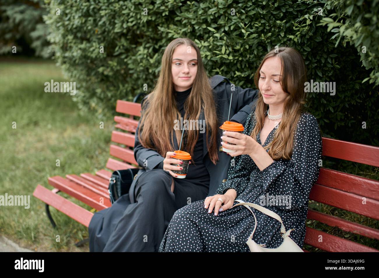Due giovani donne con lunghi capelli marroni siedono sulla panchina di legno rossa all'esterno, circondate da verde vegetale, bevono caffè e condividono momenti di relax insieme Foto Stock