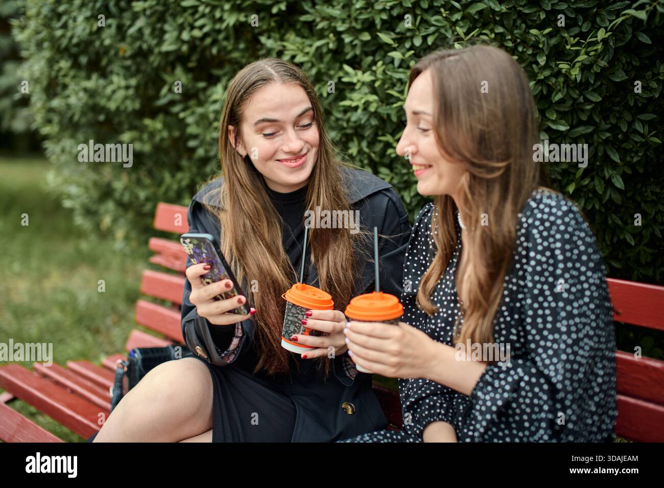 Due giovani donne adulte con capelli lunghi siedono sulla panchina rossa all'aperto, tenendo tazze da caffè da asporto e sorridendo, impegnate in pettegolezzi amichevoli con siepe verde Foto Stock