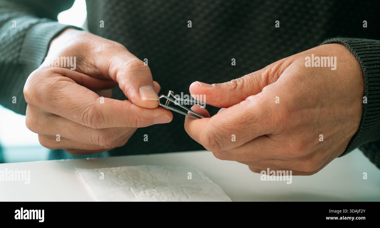 un uomo rifinisce l'unghia del pollice con un piccolo regolacapelli in metallo, il gesto è costante e preciso mentre mantiene l'igiene delle mani su una superficie bianca Foto Stock