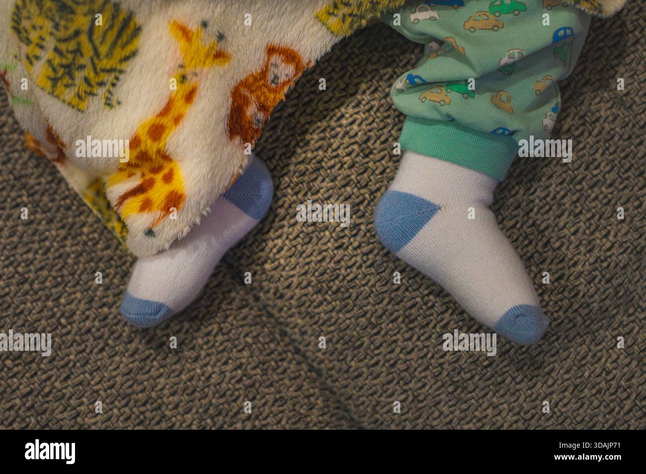 Primo piano dei piedi di un bambino in calze bianche e blu, coperte da una coperta sfocata con motivi animali, adagiate su una superficie grigia testurizzata. Foto Stock
