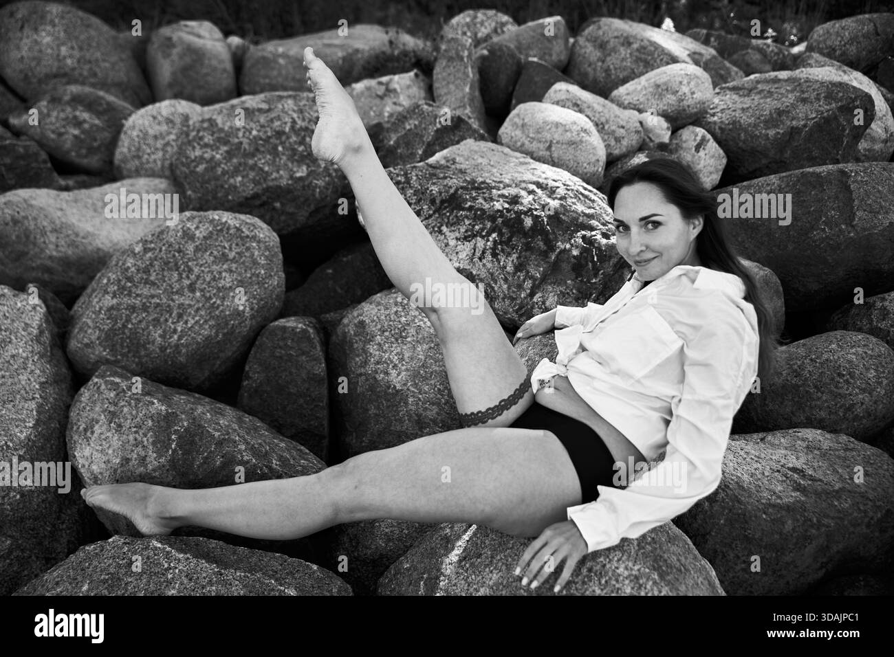 Donna adulta con i capelli scuri si inclina su grandi rocce, sollevando una gamba in alto indossando camicia bianca e pantaloni scuri. I toni monocromatici evidenziano il testo Foto Stock
