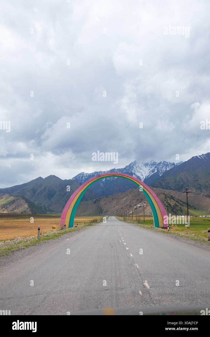 Una strada asfaltata che passa attraverso un arco colorato a forma di arcobaleno con catene montuose sullo sfondo Foto Stock