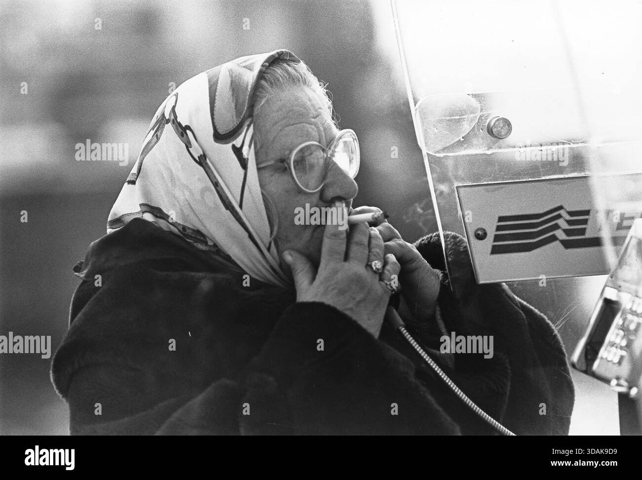 Una vecchia donna italiana sta al telefono in una cabina telefonica pubblica, fumando una sigaretta. [traduzione automatizzata] Foto Stock