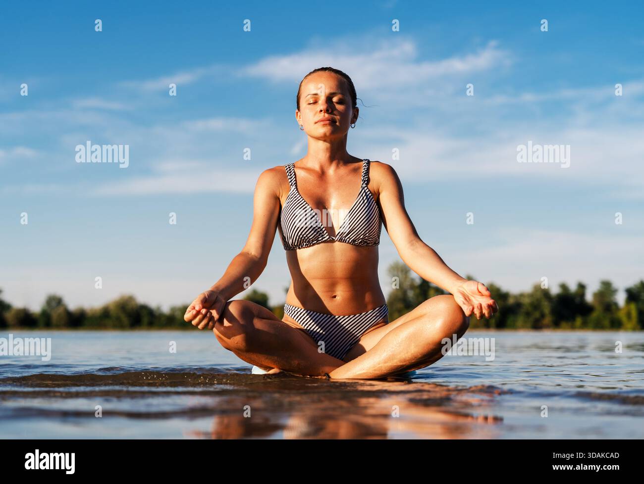 Donna serena che medita in posa di loto mentre è seduta in acque poco profonde sulla spiaggia Foto Stock