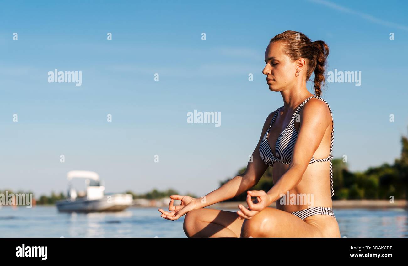 Donna serena che medita in posa di loto mentre è seduta in acque poco profonde sulla spiaggia Foto Stock