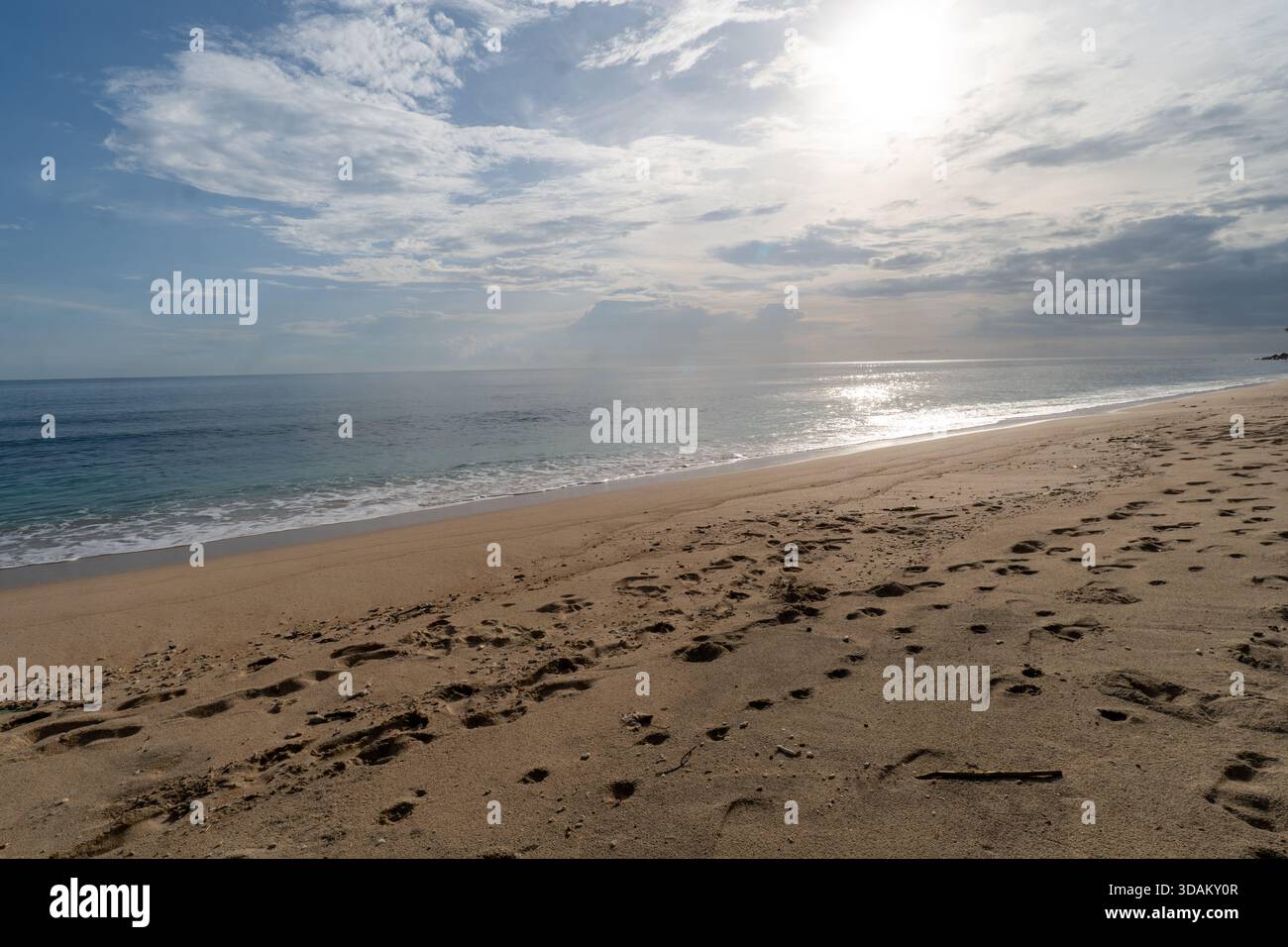 Un'ampia foto di una spiaggia sabbiosa caratterizzata da numerose impronte, onde dolci e il sole luminoso che si riflette sull'acqua dell'oceano sotto un cielo parzialmente nuvoloso. Foto Stock