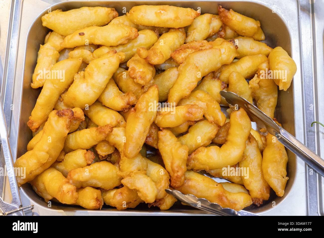 Vista ravvicinata dall'alto di una grande quantità di bastoncini di pesce pastella dorati, croccanti, impilati in un vassoio in acciaio inox, pronti per essere Foto Stock