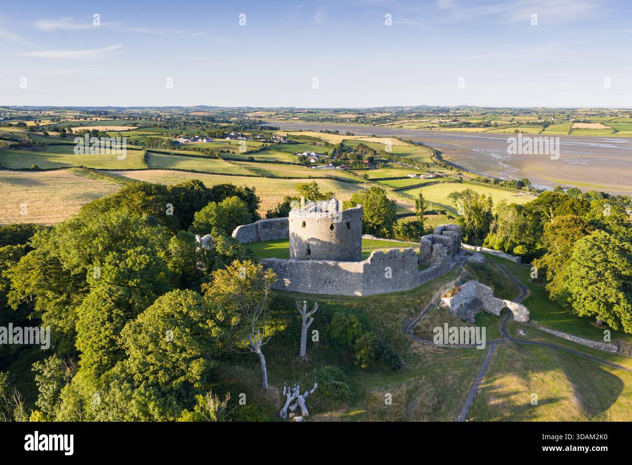 Una foto aerea panoramica di un castello medievale in rovina arroccato su una collina, circondato da lussureggianti campi, alberi e sentieri tortuosi, con una costa lontana e. Foto Stock
