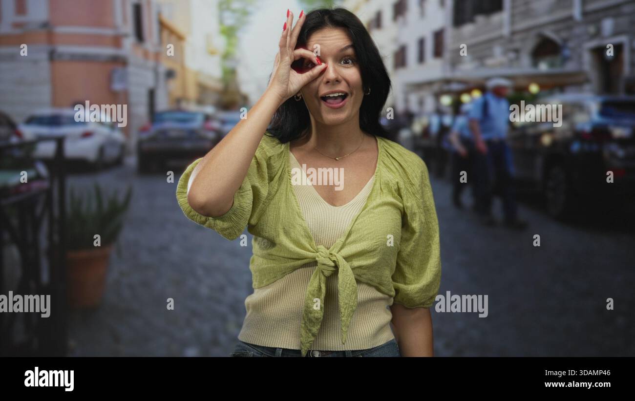 Giovane donna ispanica che fa a mano un cartello ok in strada con sopracciglia rialzata e un ampio sorriso sotto la luce del sole; consenso approvazione accordo di soddisfazione. Foto Stock