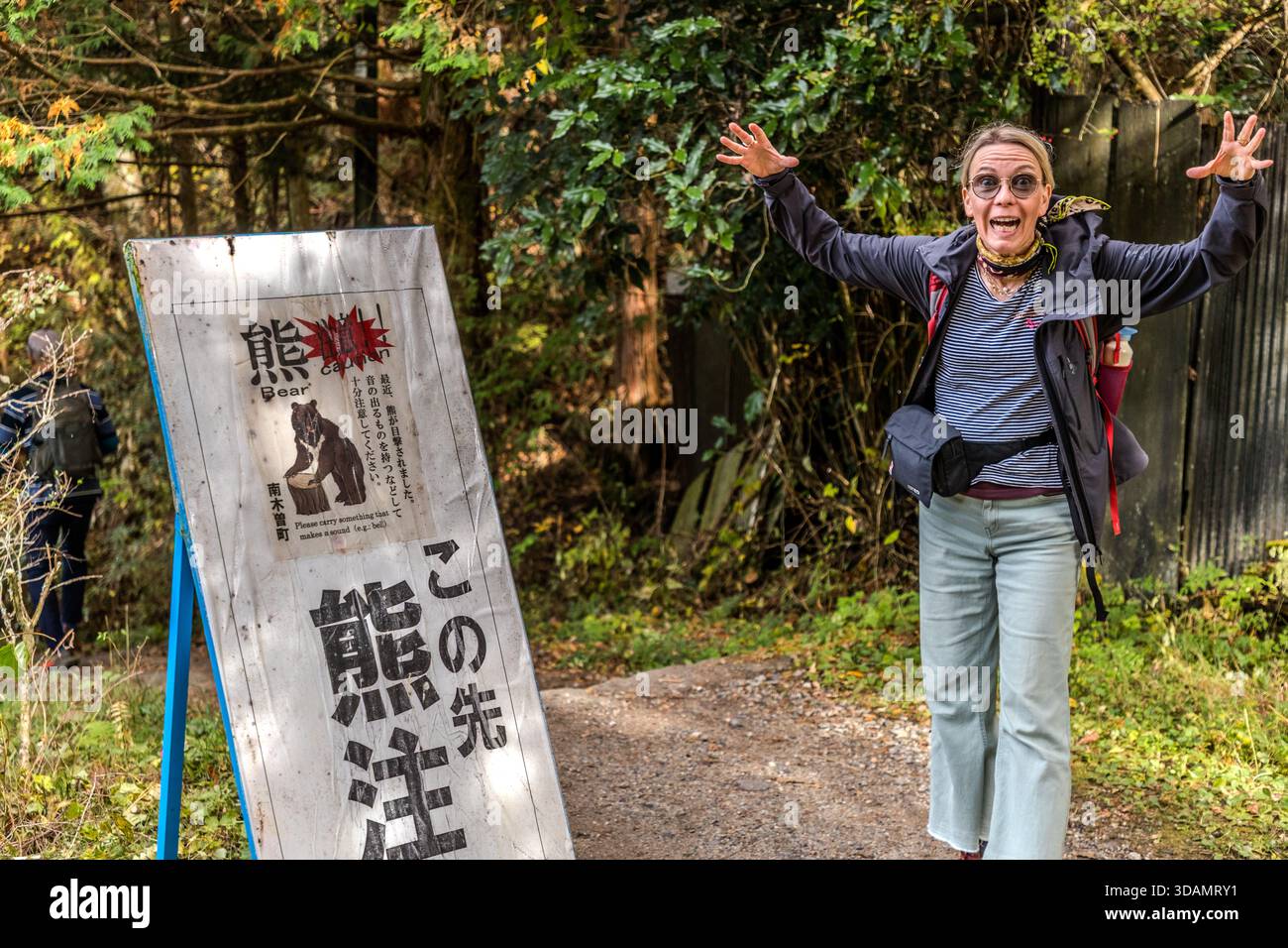 Un escursionista posa con le braccia sollevate di fronte a un cartello di avvertimento (熊注意 – «Beware of Bears») all’ingresso del sentiero forestale di Nakasendo vicino a Nagiso, prefettura di Nagano, Giappone Foto Stock