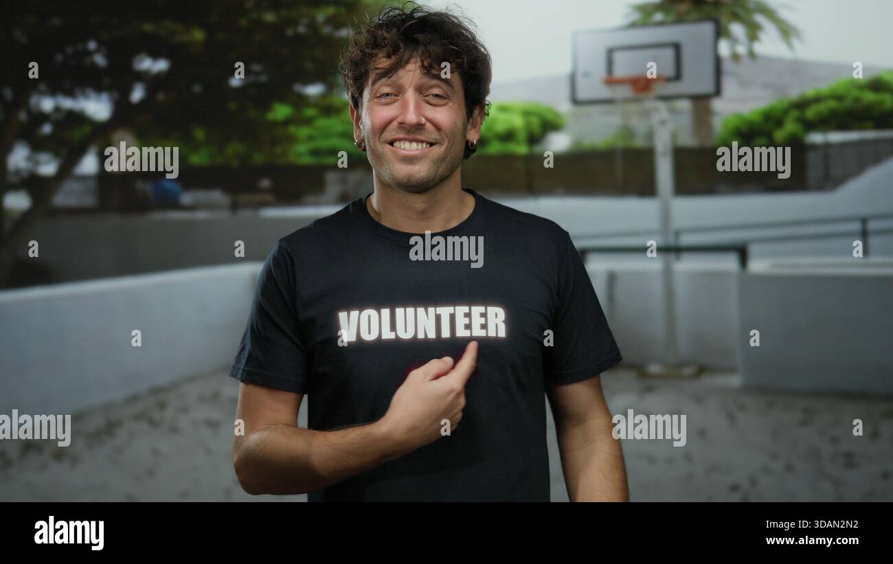 Uomo ispanico sorridente e che punta alla camicia volontaria sul campo da basket all'aperto, mostrando il coinvolgimento della comunità in un ambiente all'aperto coinvolgente. Foto Stock