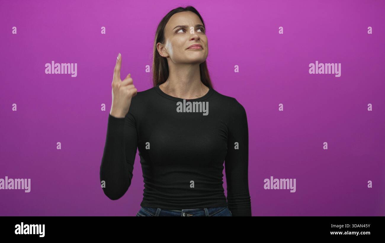La donna punta il dito indice verso l'alto nello studio magenta mentre guarda in alto e indossa camicia e jeans neri; curiosità. Foto Stock