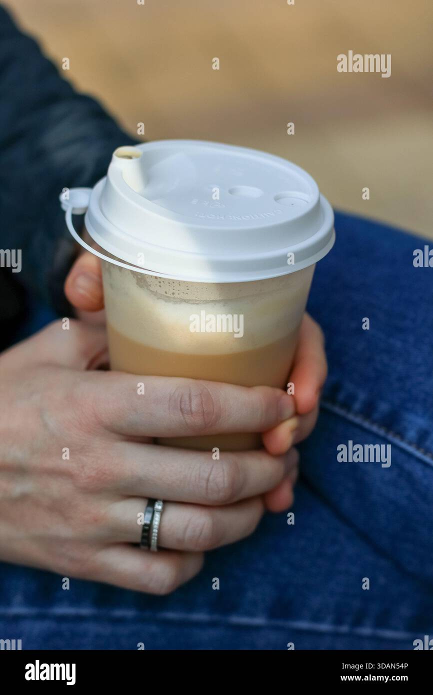 Persona in jeans che prende latte da asporto con coperchio bianco e schiuma visibile attraverso il contenitore trasparente su sfondo sfocato Foto Stock