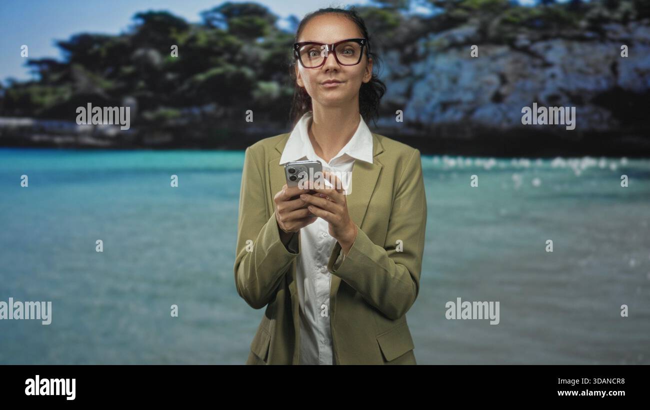 Donna con occhiali da vista, utilizza lo smartphone tenendo in mano il dispositivo e guardando in basso in uno studio con sfondo da spiaggia; concentrazione. Foto Stock